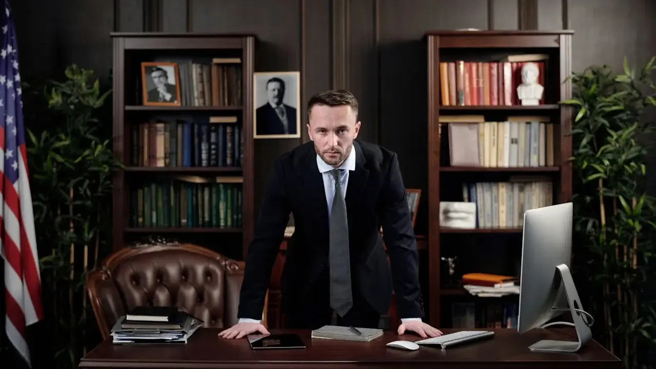 Poważny prawnik w garniturze stoi przy biurku, zastanawiając się, ile zarabia prawnik. W tle biblioteczka i amerykańska flaga.