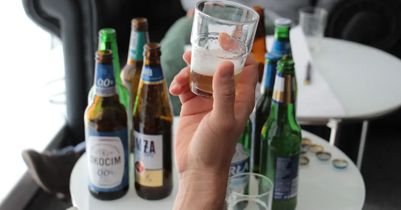 Ręka trzyma kieliszek z piwem bezalkoholowym. W tle kilka butelek piwa, idealne na przerwę w pracy.