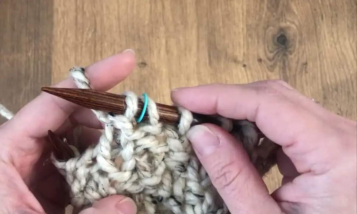 Manos tejiendo con agujas de madera un punto de relieve con lana gruesa y un marcador azul.