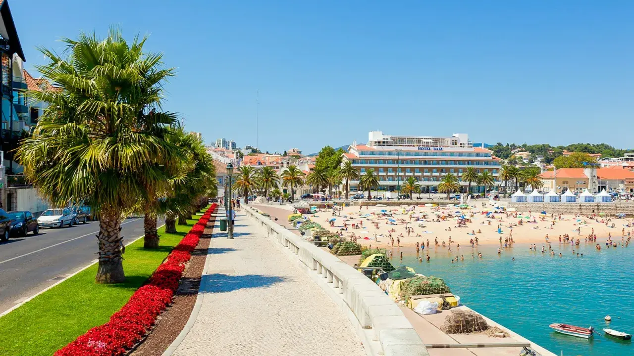 Sonniger Strandtag in Cascais bei Lissabon. Palmen säumen die Promenade, Menschen genießen das Meer und das Hotel Baía ragt empor.