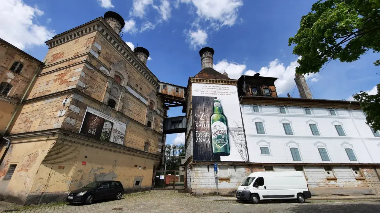 Stara fabryka z wielkimi plakatami reklamującymi piwo Namysłów. Biała furgonetka i czarny samochód stoją na brukowanej ulicy.