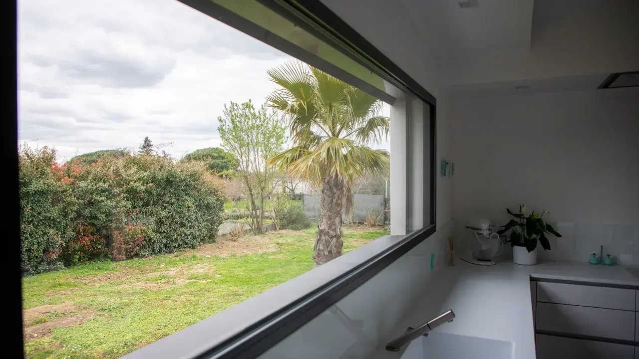Vista desde una cocina moderna con **ventanas automáticas** que enmarcan un jardín con palmera y vegetación frondosa.