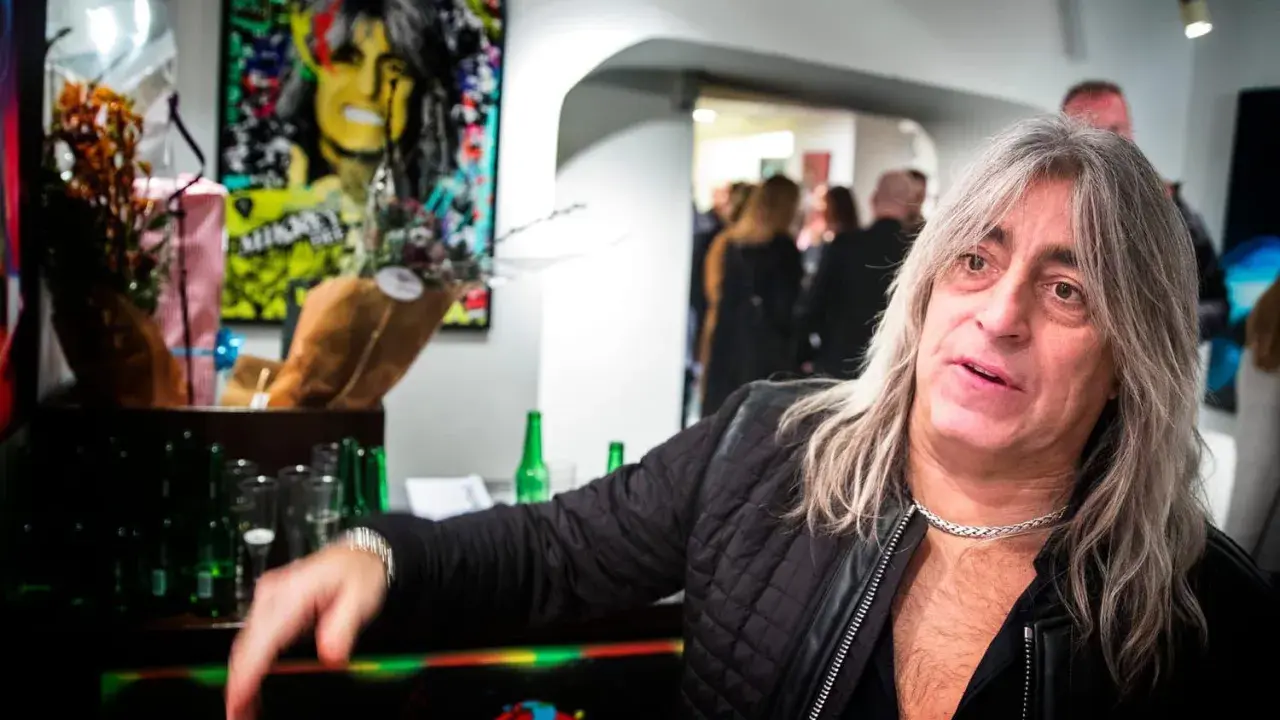 Mikkey Dee son, en man med långt grått hår och en silverfärgad kedja, poserar vid ett bord med blommor och flaskor.