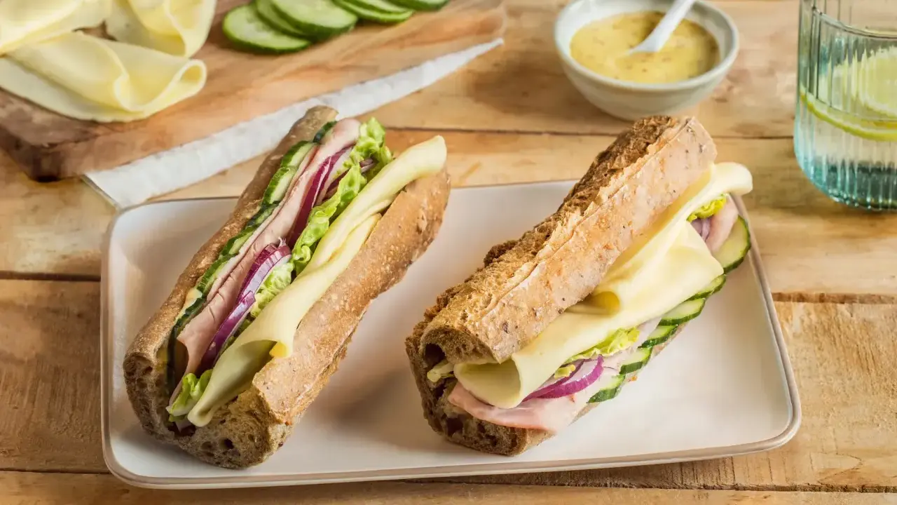 Deux sandwichs jambon fromage appétissants, garnis de concombre, oignon rouge et laitue, sur un plateau blanc.