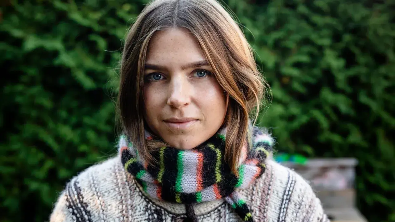 Elin Berlin med blå ögon och fräknar, klädd i en stickad tröja och randig halsduk, står framför en grön häck.