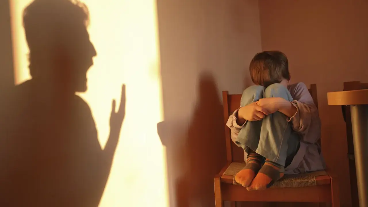 Cień krzyczącej postaci rzuca się na ścianę, podczas gdy dziecko skulone na krześle odczuwa skutki bicia dzieci w dorosłym życiu.