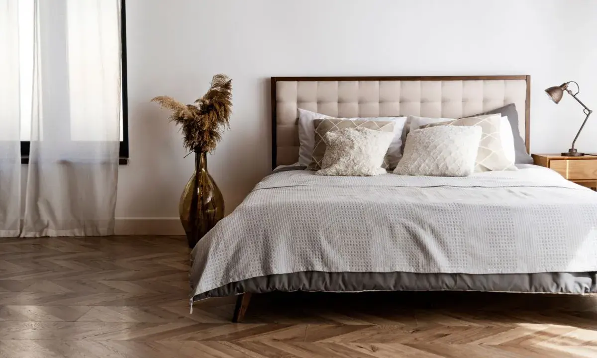 Cama con base tapizada o canapé, cojines mullidos y mantas grises. Un jarrón con pampas decora el rincón.