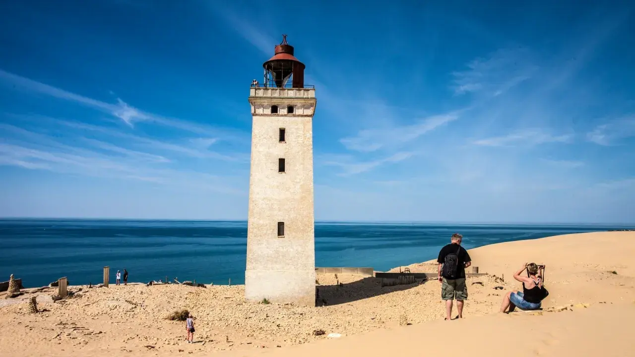 Ett vitt utsiktstorn i Danmark reser sig mot en blå himmel. Sanddyner och hav omger tornet.