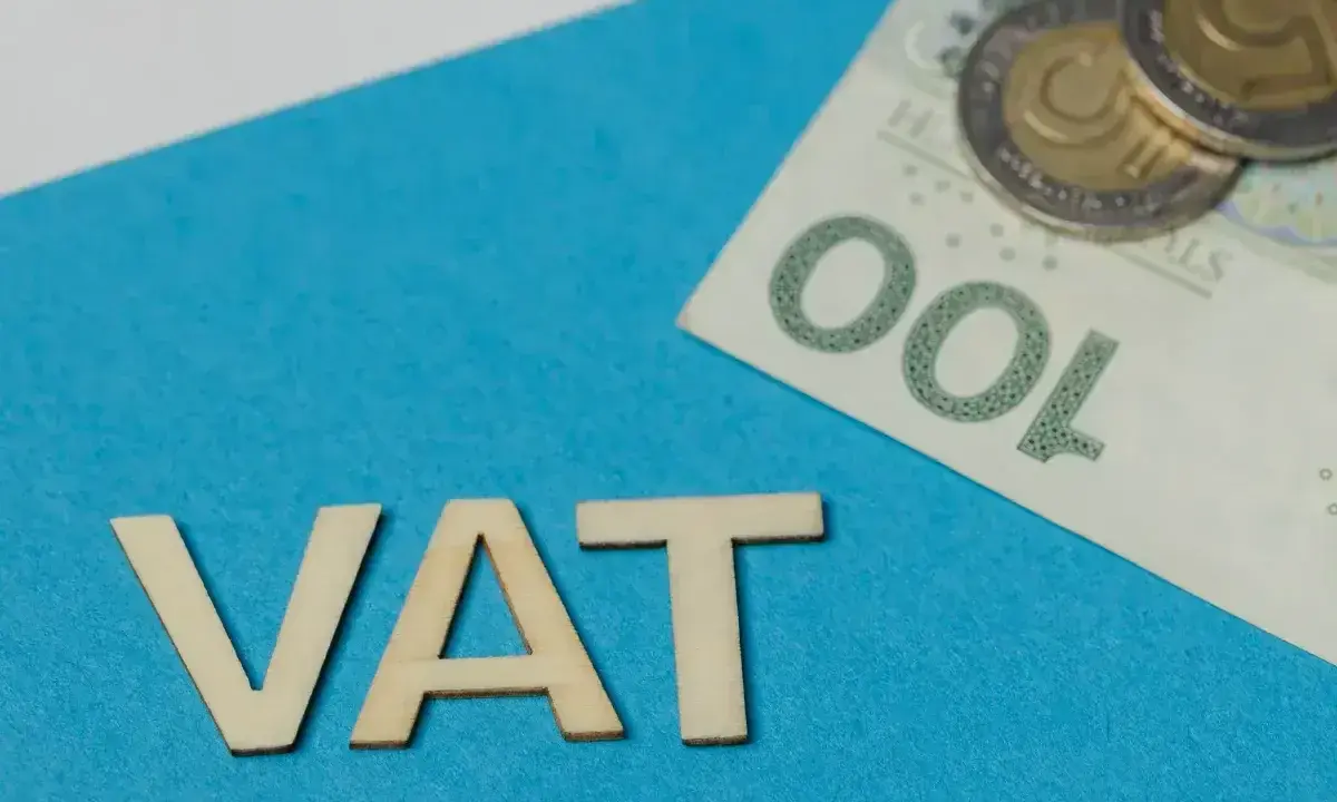 Drewniane litery VAT na niebieskim tle, obok banknot 100 zł i monety. Symbolizuje zwolnienie z VAT.