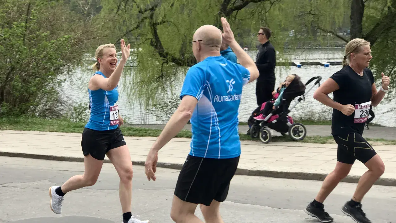Två löpare ger varandra high-five under roliga lopp. En kvinna i blått och en man i blått. I bakgrunden syns en barnvagn och en sjö.