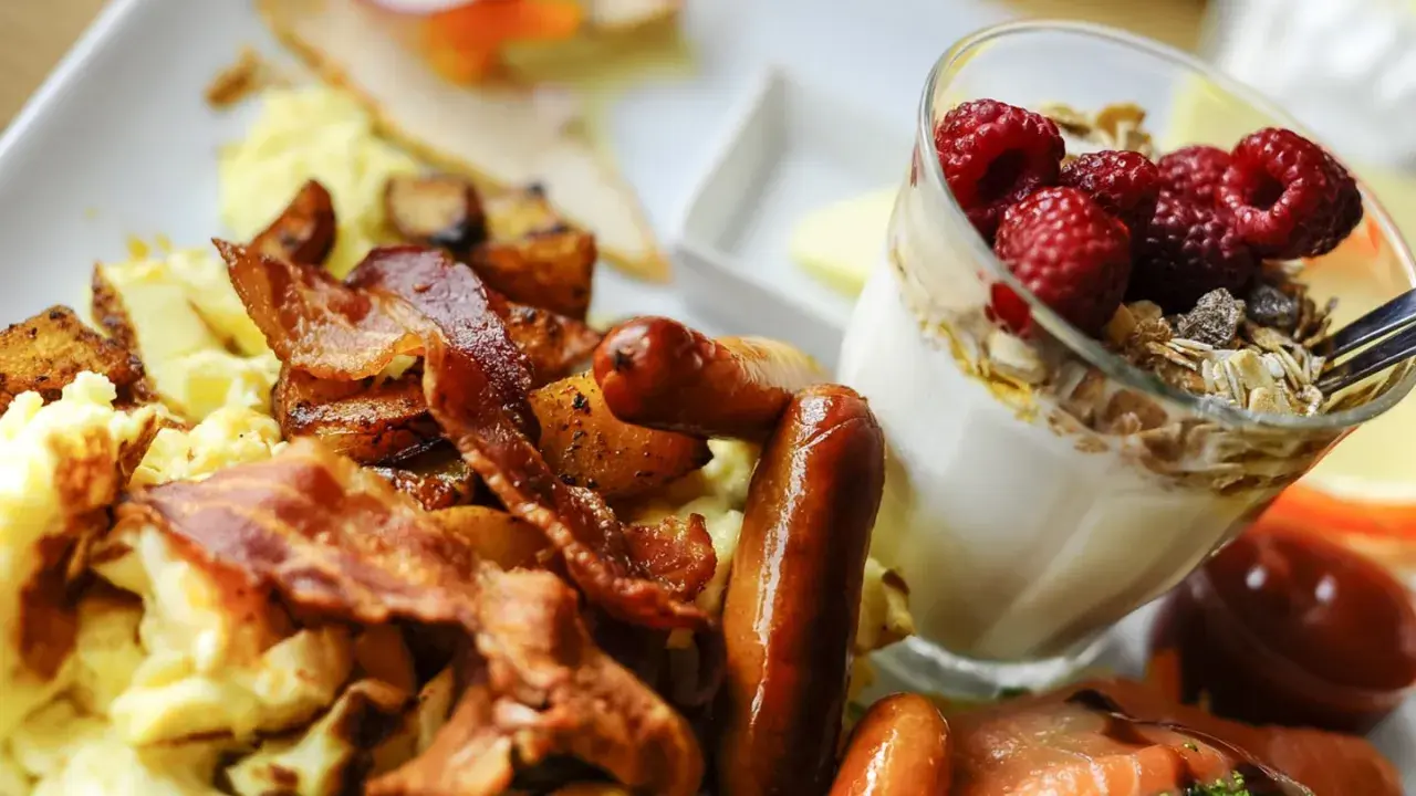 Herzhafte Ideen zum Brunch: Rührei, Speck, Würstchen und Kartoffeln neben einem Glas Joghurt mit Beeren und Müsli.