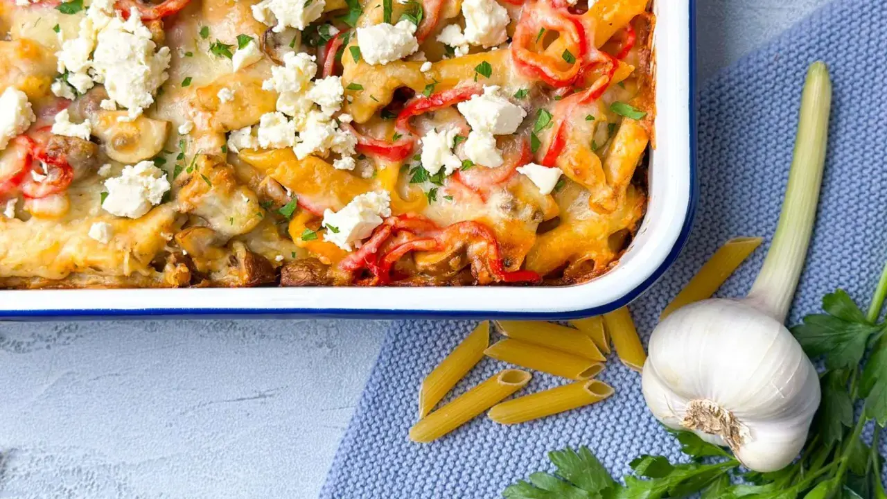 Herzhafter Gyros Auflauf mit Penne, Paprika und Feta, garniert mit Petersilie. Daneben liegen Penne, Knoblauch und frische Kräuter.