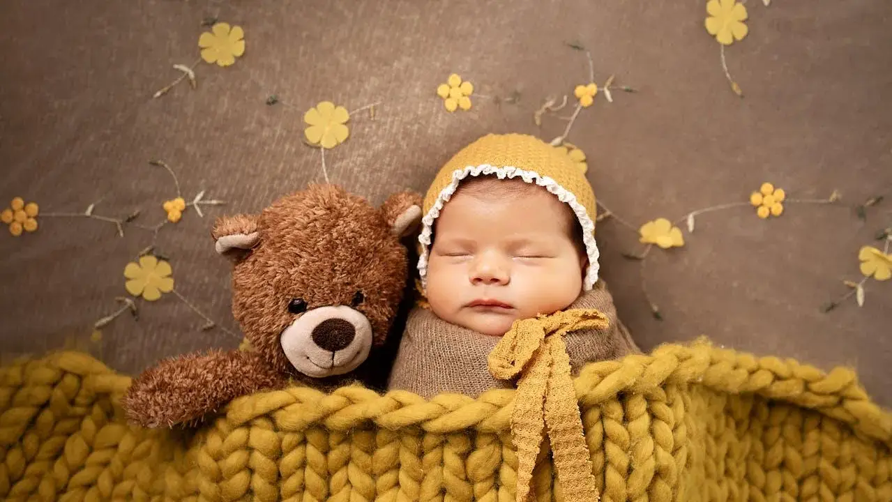 Bambino addormentato con orsacchiotto, avvolto in una coperta gialla. Un'idea per foto neonati fai da te con decorazioni floreali.
