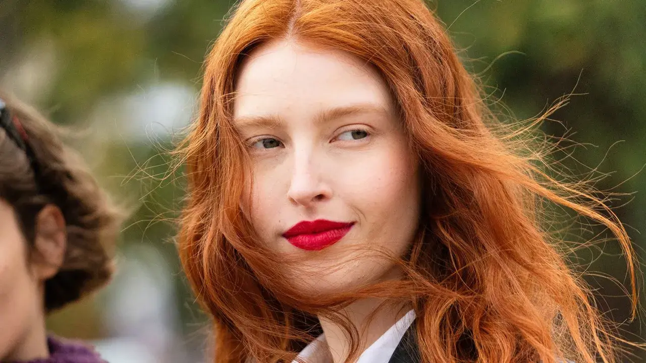 Eine Frau mit leuchtend roter haarfärbung und roten Lippen, ihr Haar weht im Wind.