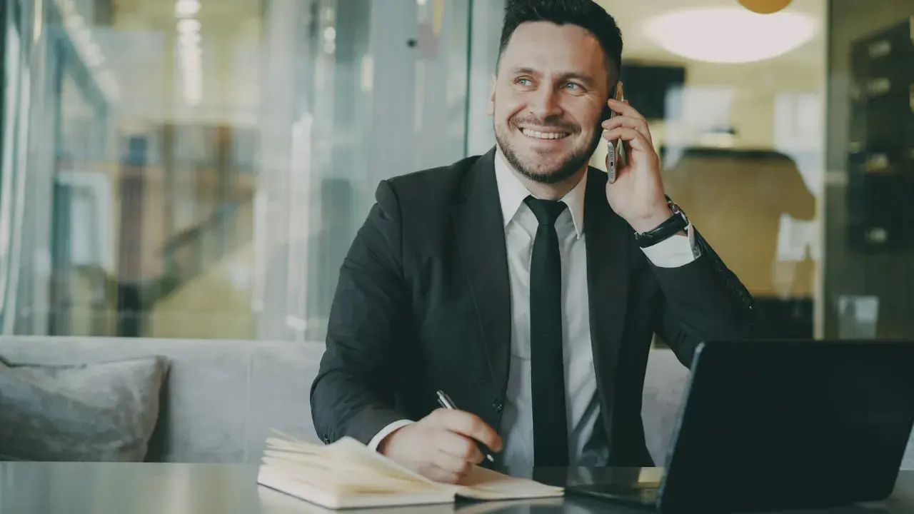 Uśmiechnięty mężczyzna w garniturze rozmawia przez telefon, notując coś w zeszycie. To właśnie taki mikroprzedsiębiorca buduje swój biznes.