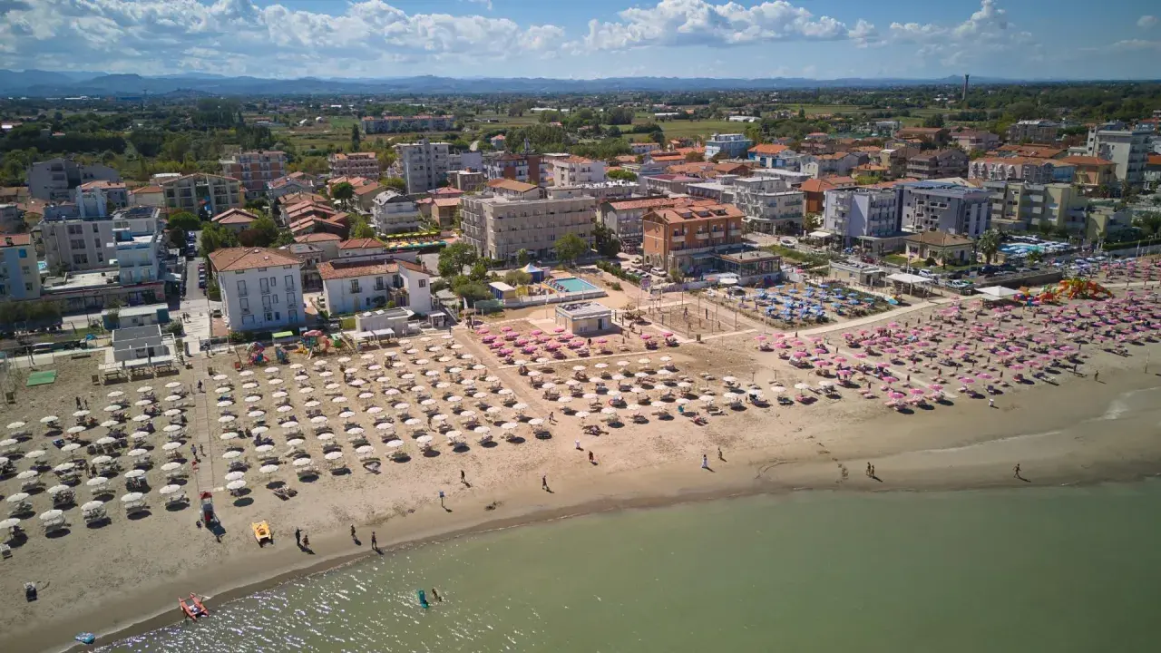 Vista aerea di una spiaggia affollata con ombrelloni rosa e bianchi, hotel e case nel centro storico di Torre Pedrera.