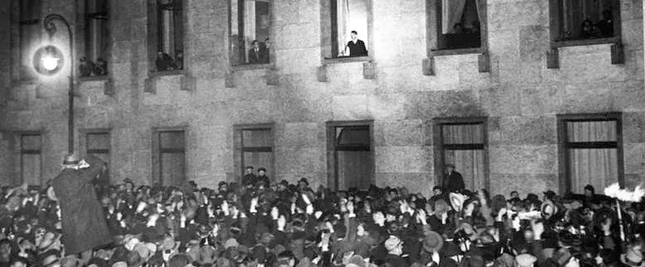 Menge jubelt vor Gebäude, während ein Mann auf einer Leiter steht. Die Szene erinnert an die Zeit des Ermächtigungsgesetzes.