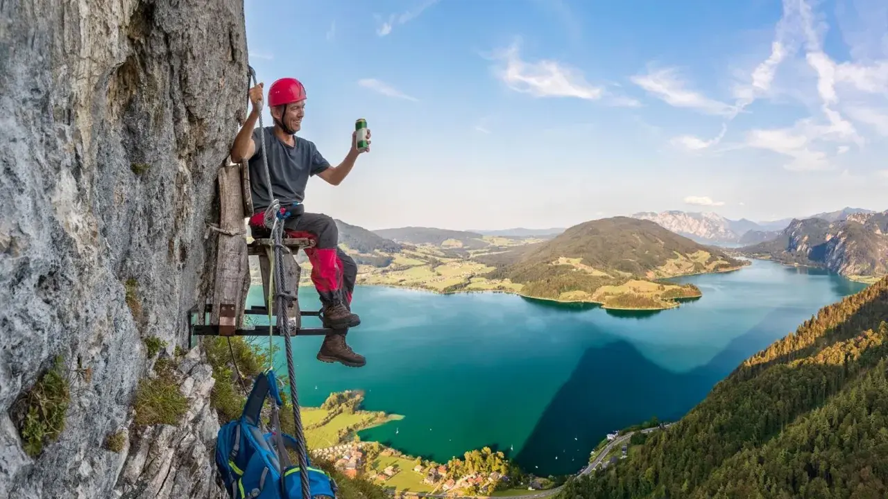 Mężczyzna w kasku siedzi na drewnianej platformie przy skalnej ścianie, ciesząc się widokiem na jezioro w Austrii. To wspaniała przygoda na via ferrata Austria!
