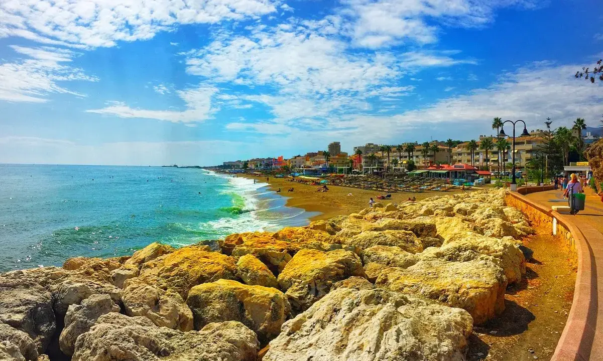 Sonniger Tag an der playa de la carihuela. Türkises Meer, goldener Sandstrand mit Liegen und Palmen, gesäumt von bunten Gebäuden. Felsen im Vordergrund.