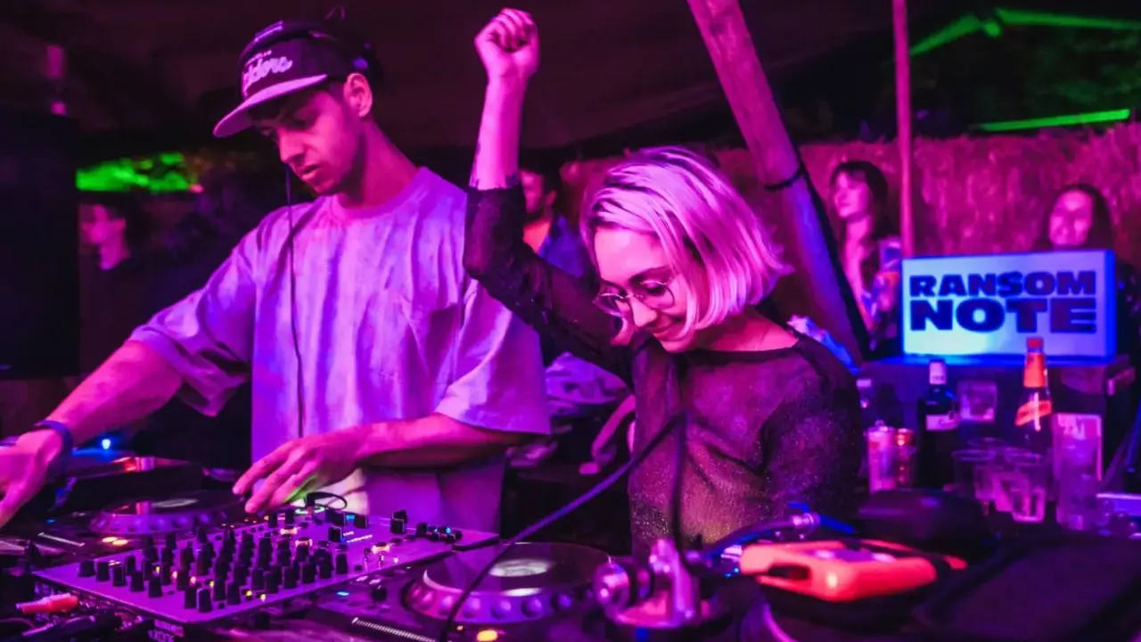 DJ set b2b: un uomo e una donna mixano musica in un locale con luci viola e verdi. La folla balla.