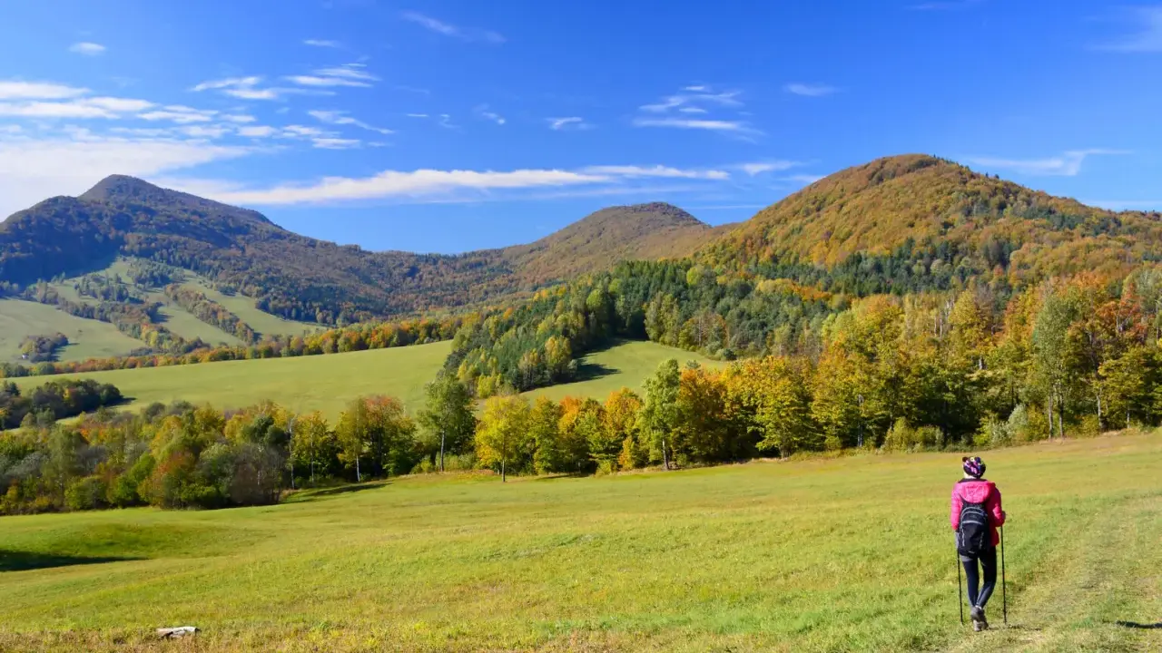 Jesień na **Beskid Niski szlaki**. Turystka w różowej kurtce idzie przez zieloną łąkę w kierunku kolorowych wzgórz.