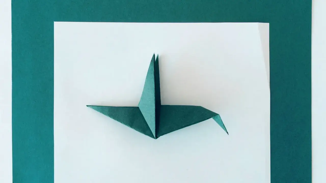 Zielony żuraw z papieru, przykład sztuki origami dla dorosłych, leży na białym tle otoczonym ciemnozielonymi ramkami.