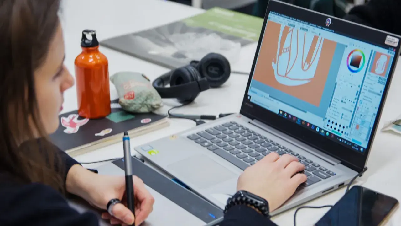 Studentka pracuje nad projektem w **studiach graficznych Warszawa**. Na ekranie laptopa widać program do grafiki, a obok słuchawki i butelkę z wodą.