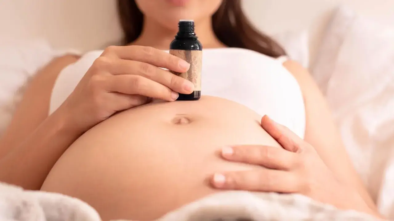 Schwangerenbauch mit einer Hand, die eine kleine Flasche Öl hält. Ab wann Dammmassage?