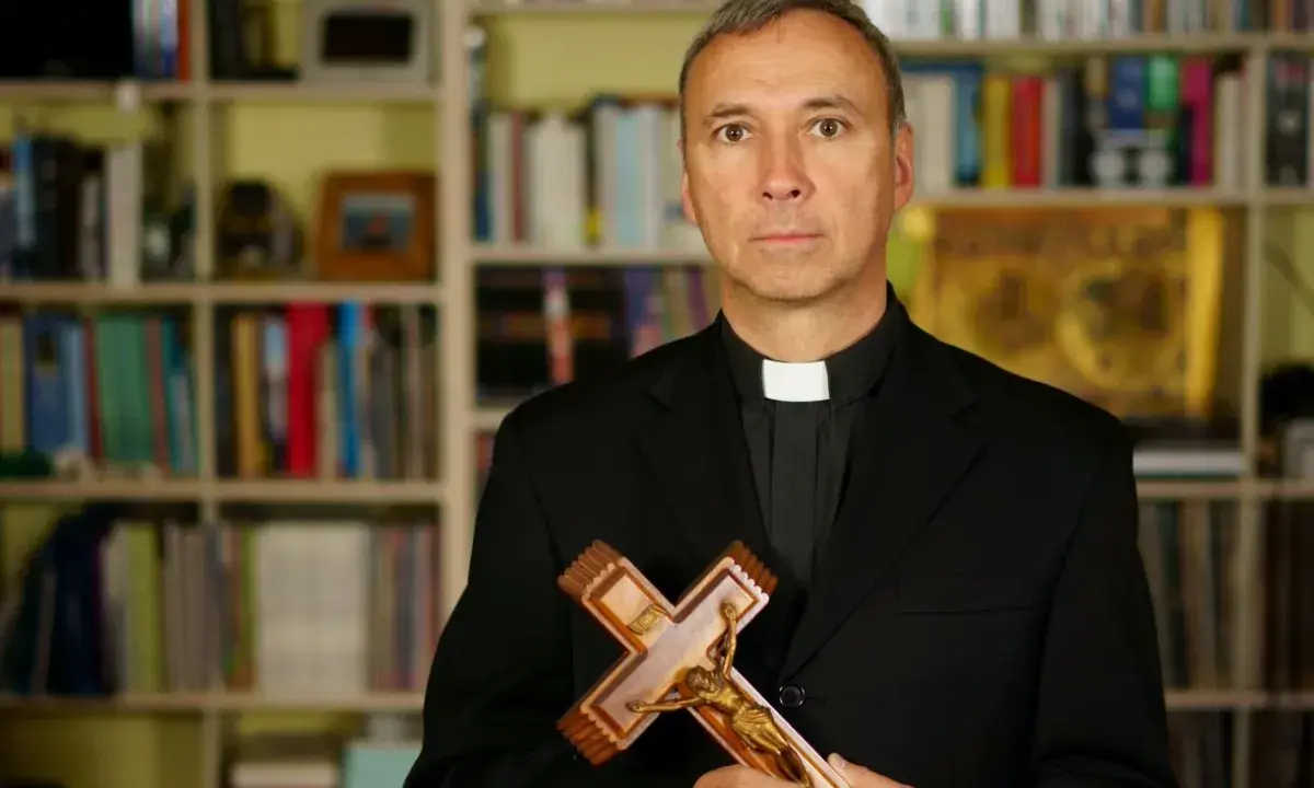 Duchowny z krzyżem w dłoniach, na tle regałów z książkami. Czy religia wlicza się do czerwonego paska?