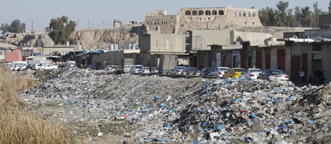 Müllberge säumen die Straßen von Kirkuk, Irak. Autos parken hinter den Geschäften, während im Hintergrund alte Gebäude auf einem Hügel stehen.
