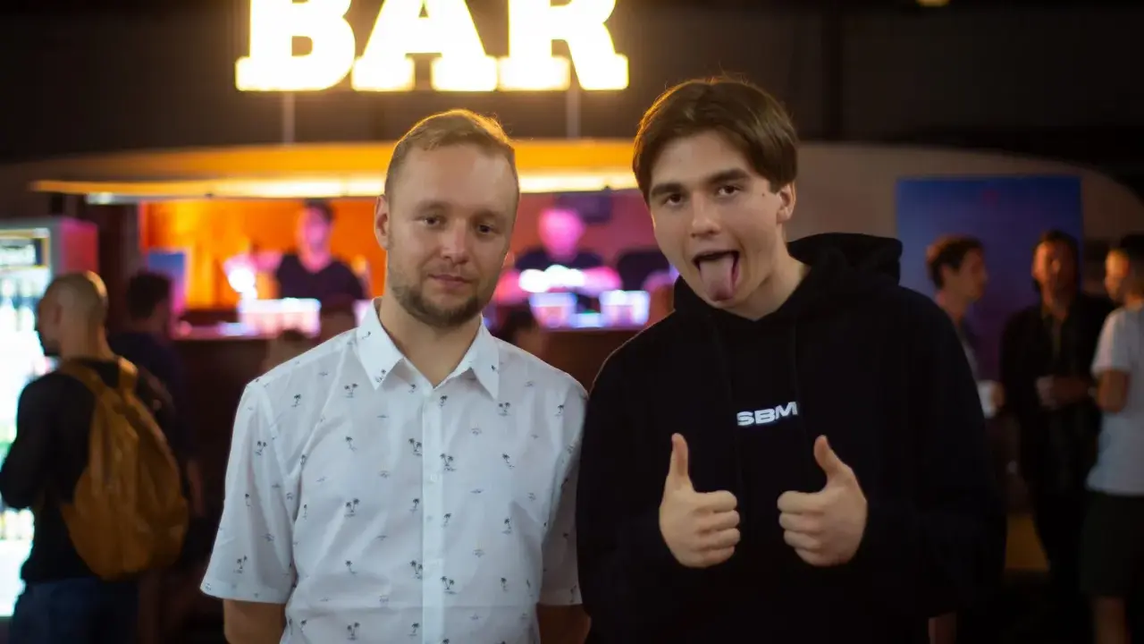 Dwóch mężczyzn pozuje przed napisem "BAR". Jeden w koszuli w palmy, drugi w bluzie z napisem SBM, pokazuje kciuki w górę i wystawia język.