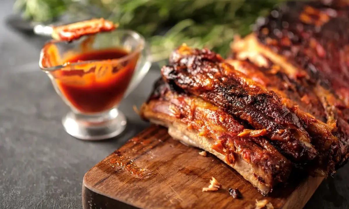 Costine al forno succulente e caramellate, servite su tagliere di legno con salsa barbecue in ciotolina.