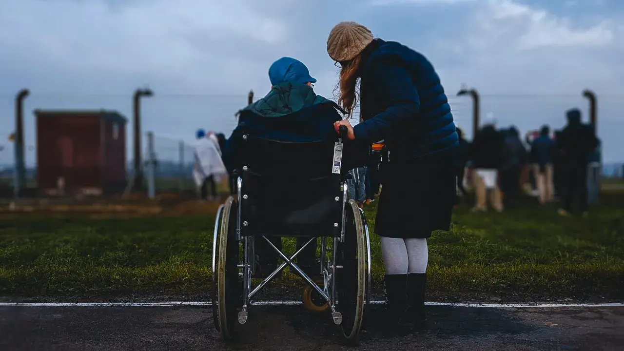 Accompagnamento anziani a chi spetta: una donna spinge una persona in sedia a rotelle, con altre figure sfocate sullo sfondo.