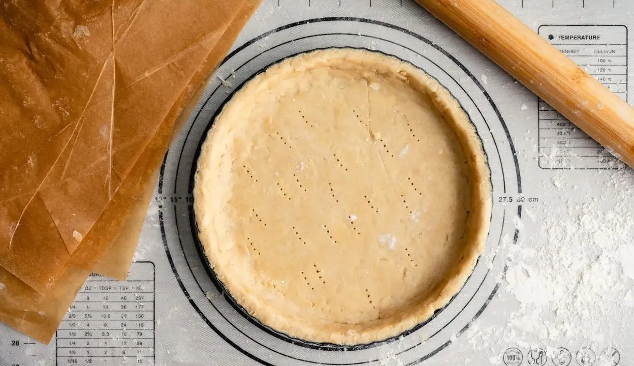 Pâte sucrée noisette prête à être garnie, sur un tapis de pâtisserie avec un rouleau à bois et du papier sulfurisé.