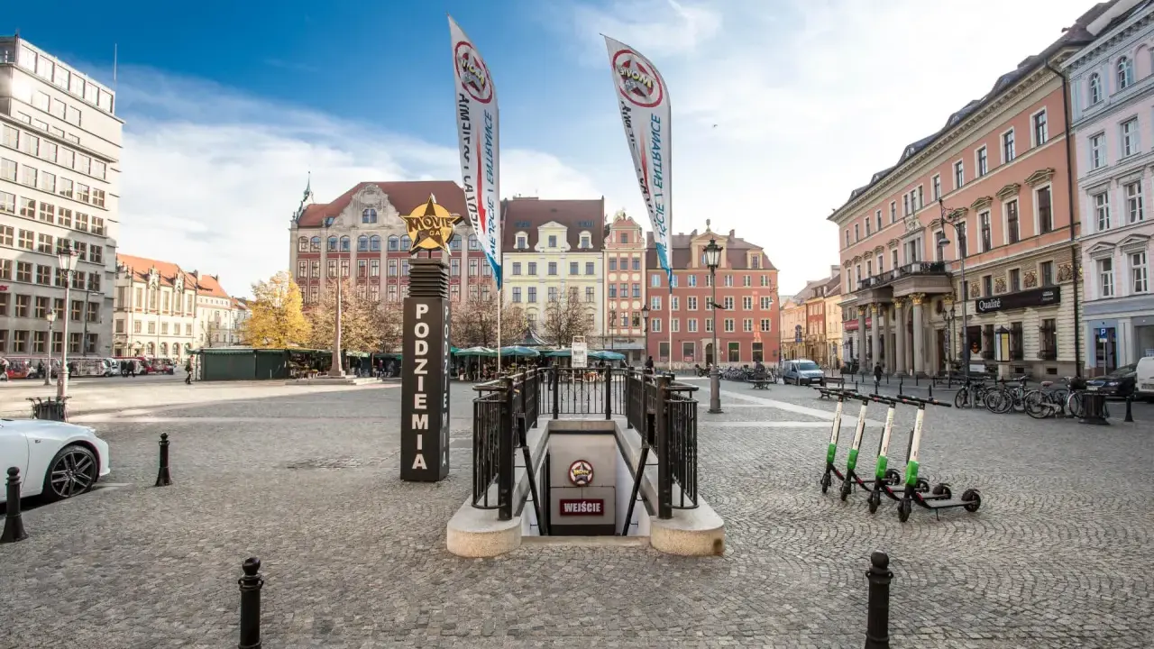Wejście do podziemi z napisem "Movie Gate" i dwoma pionowymi banerami. W tle kolorowe kamienice i błękitne niebo.
