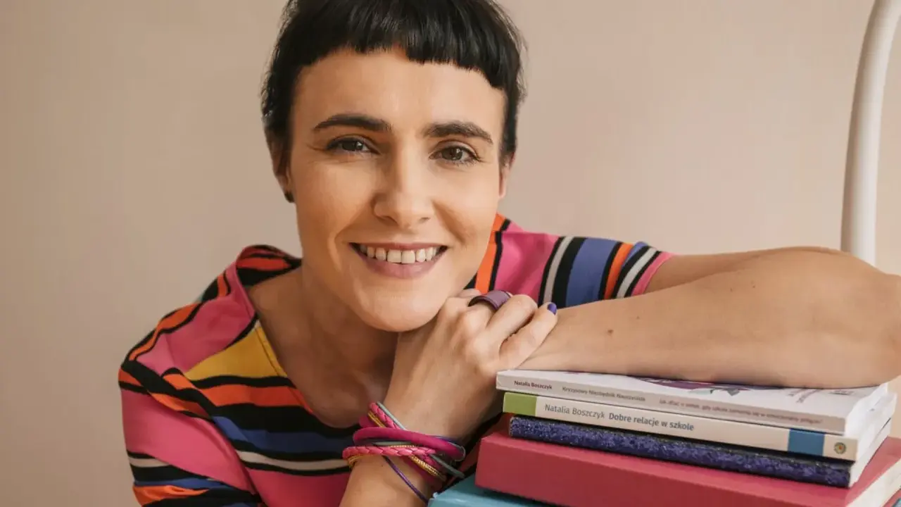 Uśmiechnięta kobieta z krótkimi włosami opiera się o stos kolorowych książek. Wiedza, którą posiadają, jest dla niej dostateczna.