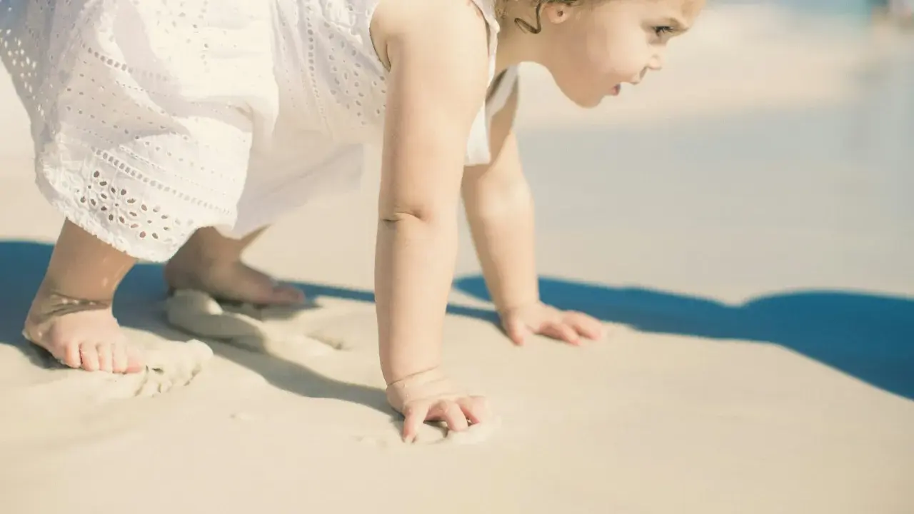Małe dziecko w białej sukience raczkuje po plaży. Może to być jedna z przyczyn nie chodzenia dziecka, bo piasek jest ciekawy.