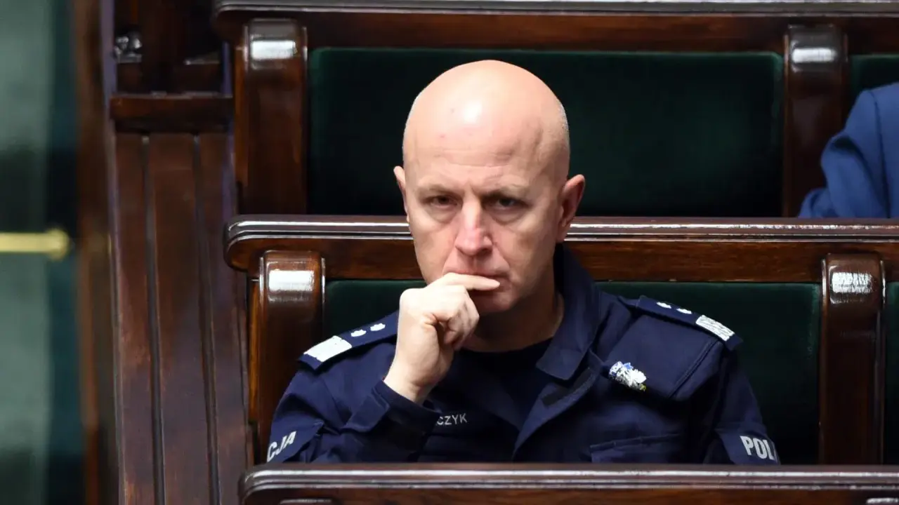 Łysy mężczyzna w mundurze policji siedzi w drewnianej ławie, zamyślony.