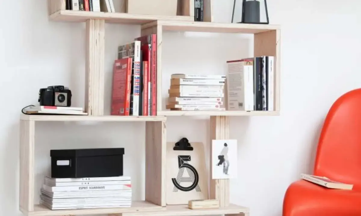 Bibliothèque DIY en bois clair, remplie de livres, d'une caméra vintage et d'une boîte noire. Une chaise rouge vif est visible à droite.