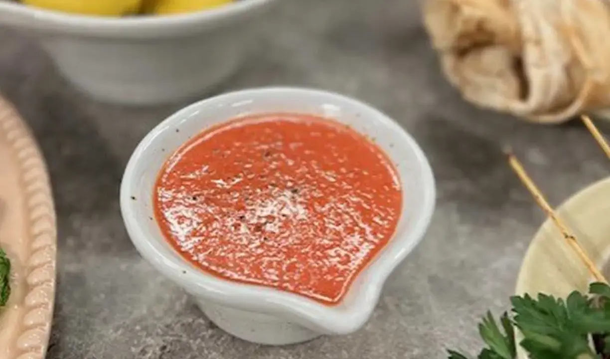 En skål med en röd, kryddig sås, kanske en pico de gallo, står på ett grått bord. Bredvid syns gula citroner och bröd.