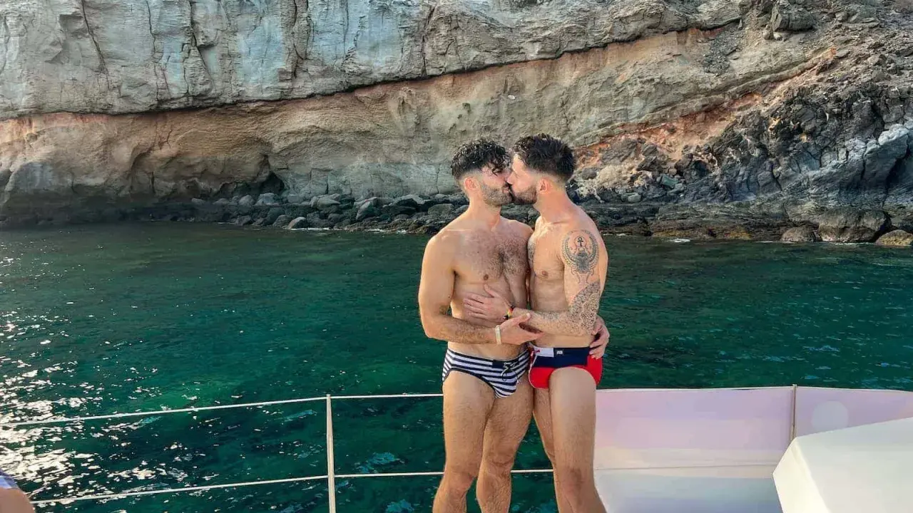 A loving gay couple shares a kiss on a boat, enjoying the stunning scenery of the Canary Islands.