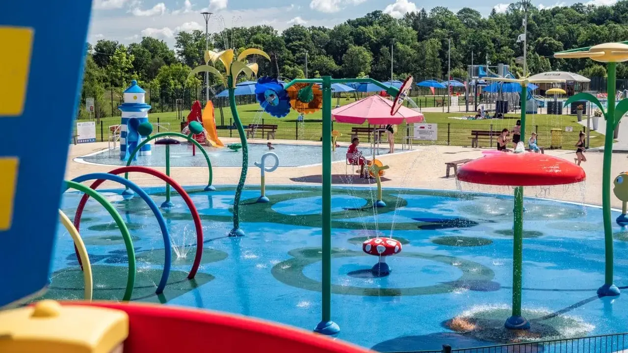 Kolorowy wodny plac zabaw z atrakcjami dla dzieci w Krosnie. Dzieci pluskają się w wodzie pod kolorowymi parasolami i zraszczaczami.