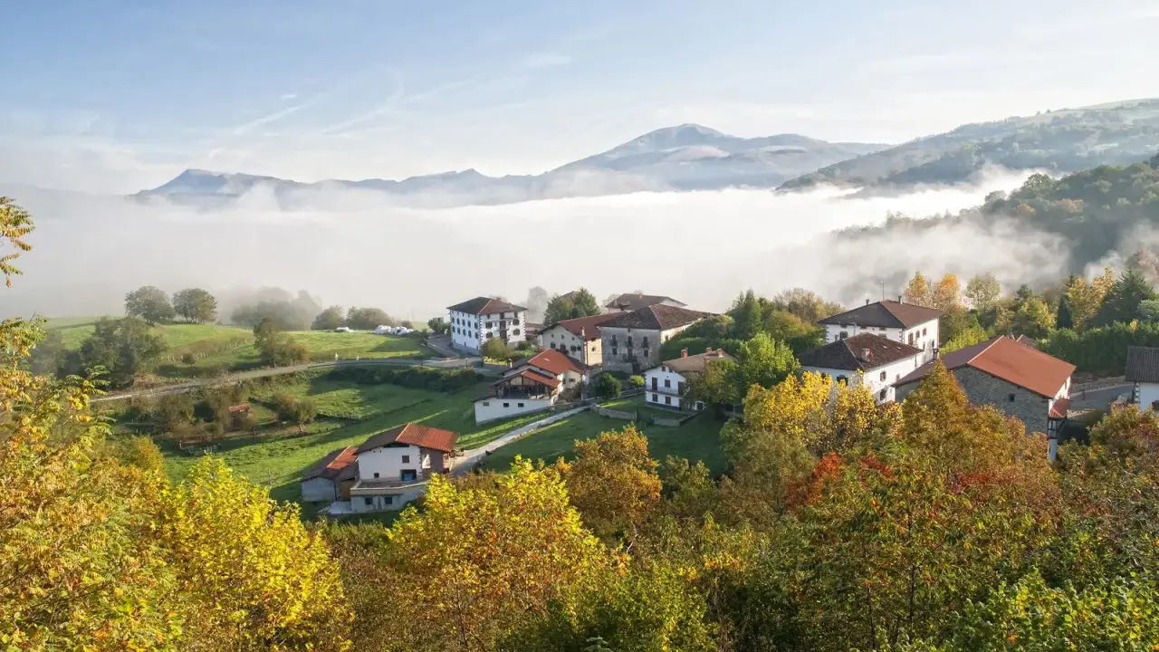 Pueblo en el **valle del Baztan** cubierto por niebla matutina, con casas blancas y tejados rojos entre árboles otoñales.