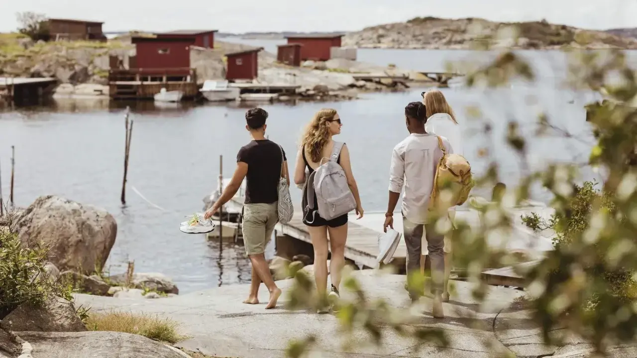 Vänner njuter av en promenad längs vattnet vid röda fiskestugor, en perfekt bild av eco tourism i Sverige.