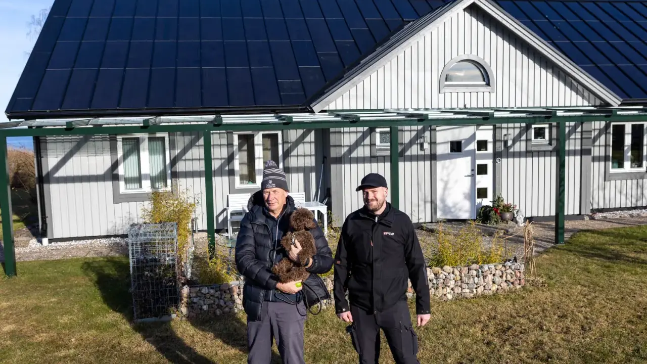 Två män och en hund framför ett hus med solpaneler på taket. Taket har vackra vinklar som ger huset karaktär.
