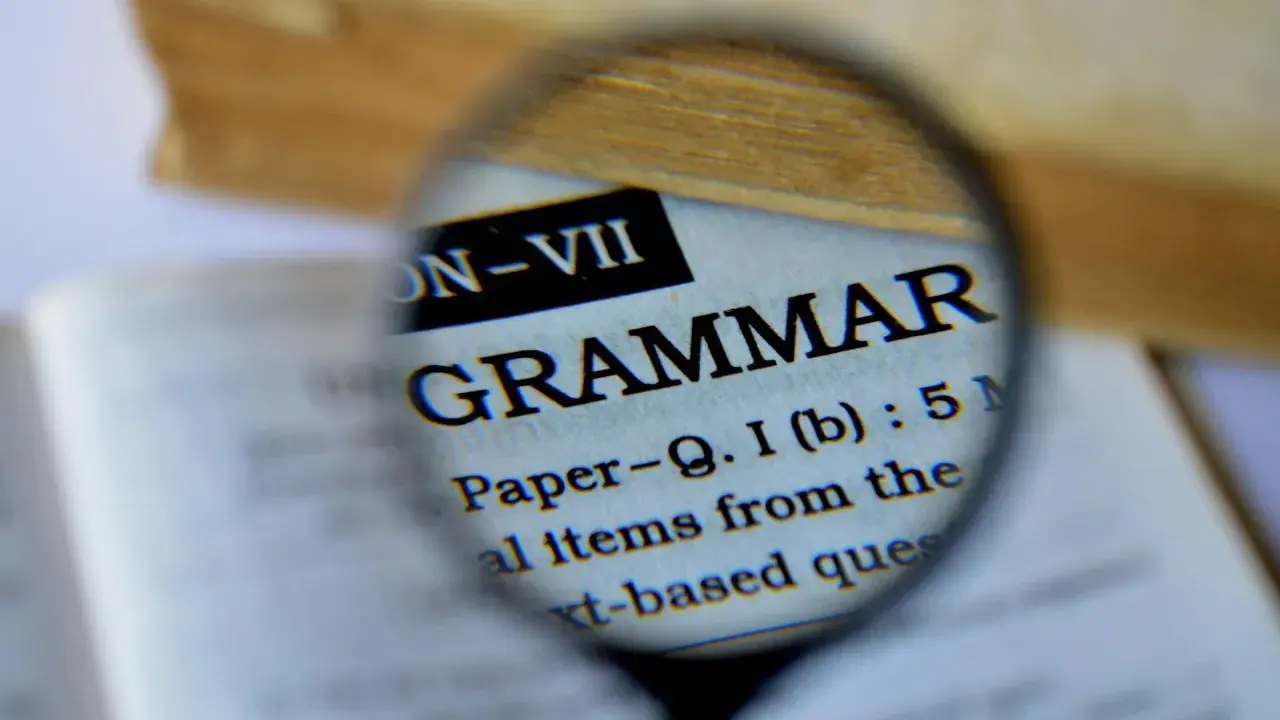 Une loupe examine le mot "GRAMMAR" dans un livre. Pour maîtriser la langue, il faut bien receler la conjugaison.