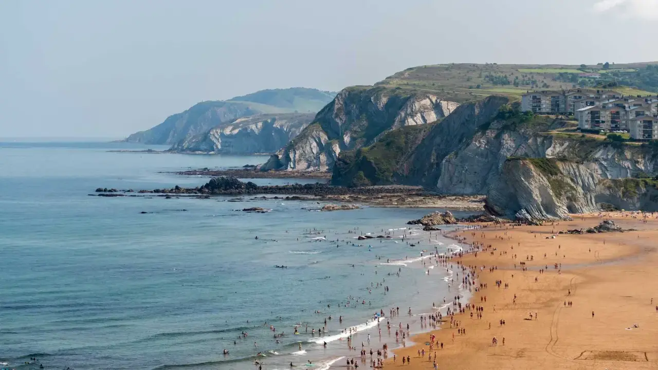 Ein belebter Strand mit vielen Menschen, die im Meer schwimmen und surfen, vor einer dramatischen Felsküste. Das Bilbao Meer lädt zum Entspannen ein.