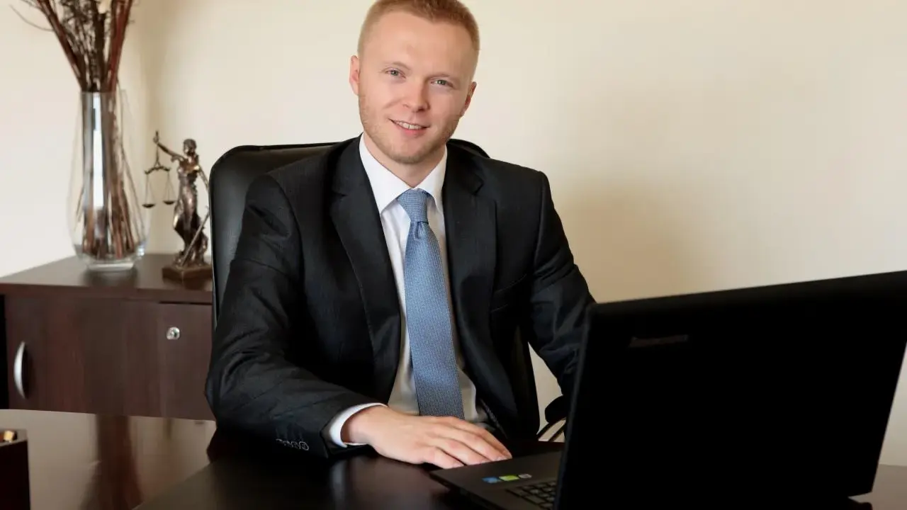 Mężczyzna w garniturze siedzi przy biurku z laptopem, obok statuetka Temidy.