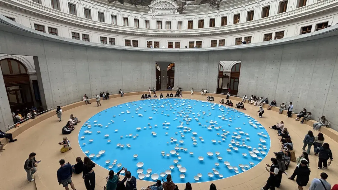 A circular room with a frescoed ceiling, featuring a bright blue pool dotted with white ceramic bowls. Visitors observe this installation, part of the François-Henri Pinault art collection.