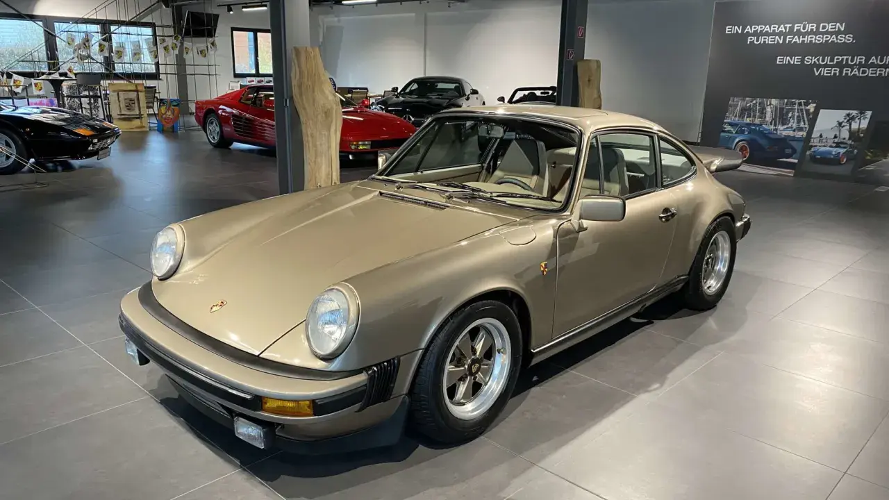 Ein goldener Porsche 911 SC 3.0 steht in einer Ausstellungshalle, flankiert von anderen Sportwagen.